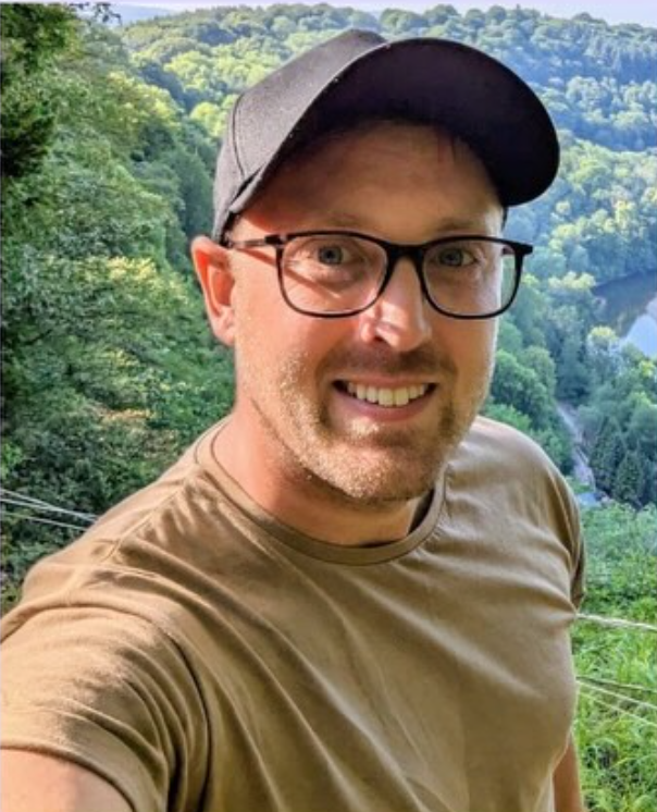 Alex Green smiling outdoors, wearing glasses and a cap